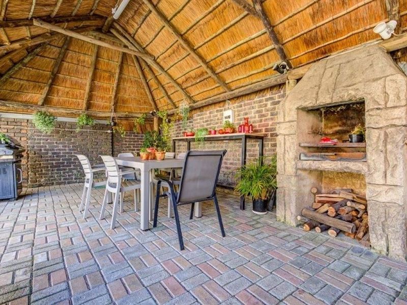 Covered outdoor patio featuring a traditional thatch roof, exposed brick walls, and a built-in stone braai area, perfect for outdoor entertaining.