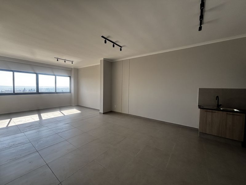 Spacious open-plan living area featuring modern tiled flooring, track lighting, and a compact kitchenette with a sink, offering bright natural light.