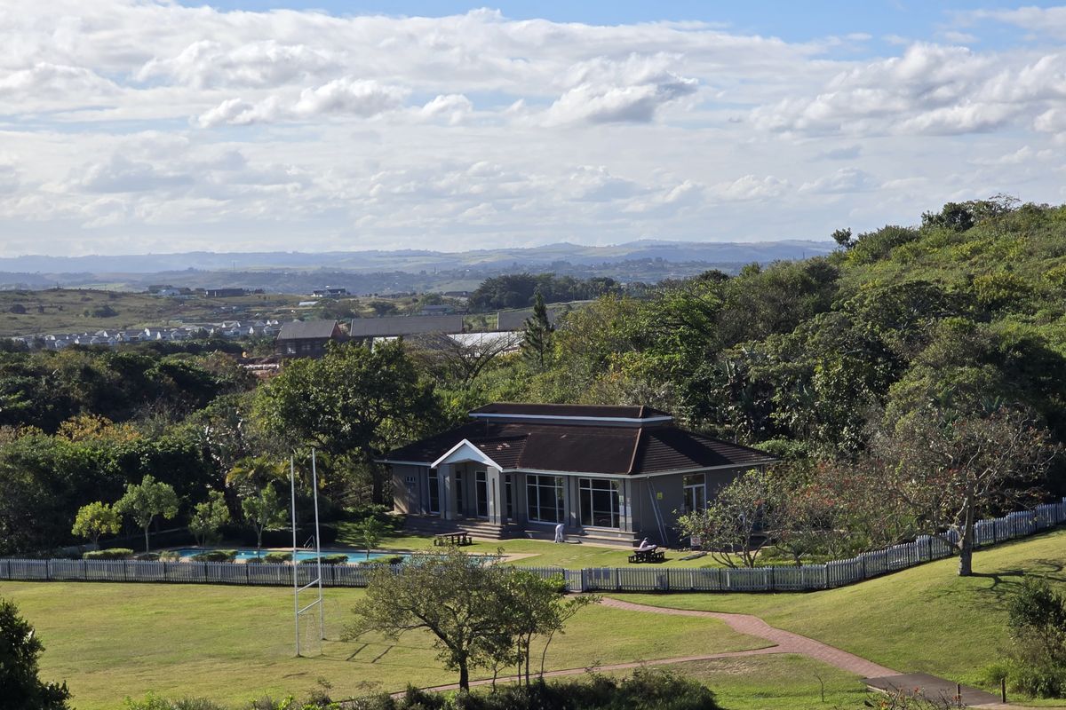 An elevated aerial view of a clubhouse facility featuring a swimming pool, landscaped grounds, and a picket fence, set within a scenic estate.