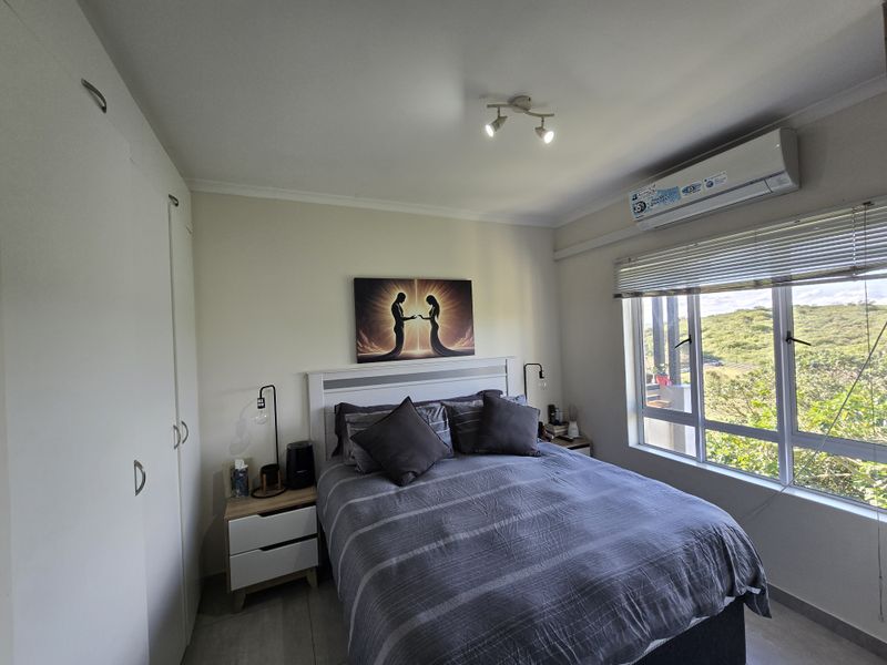 A well-maintained bedroom featuring built-in wardrobes, a split-system air conditioner, and large windows with venetian blinds offering natural light and scenic views.