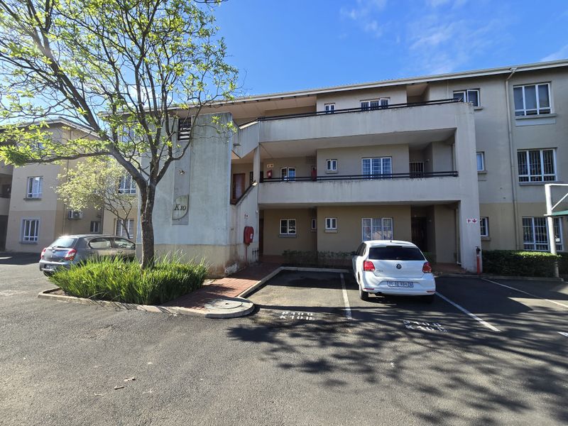 Front exterior view of a multi-story apartment complex featuring paved parking bays, balconies, and shared access walkways.