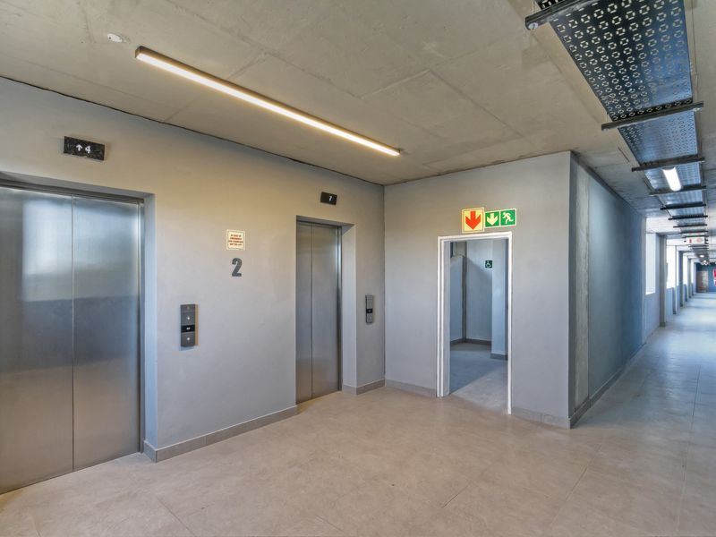 A modern apartment building hallway featuring elevator access, exposed concrete ceilings, and tiled flooring with clear emergency exit signage.