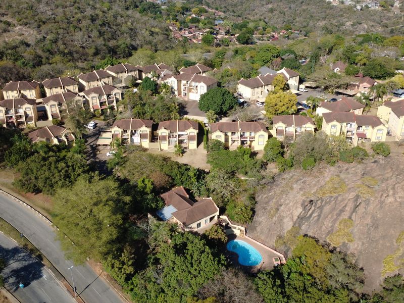 Aerial view of a residential apartment complex featuring multiple buildings, lush greenery, and a private swimming pool set against a rocky landscape.