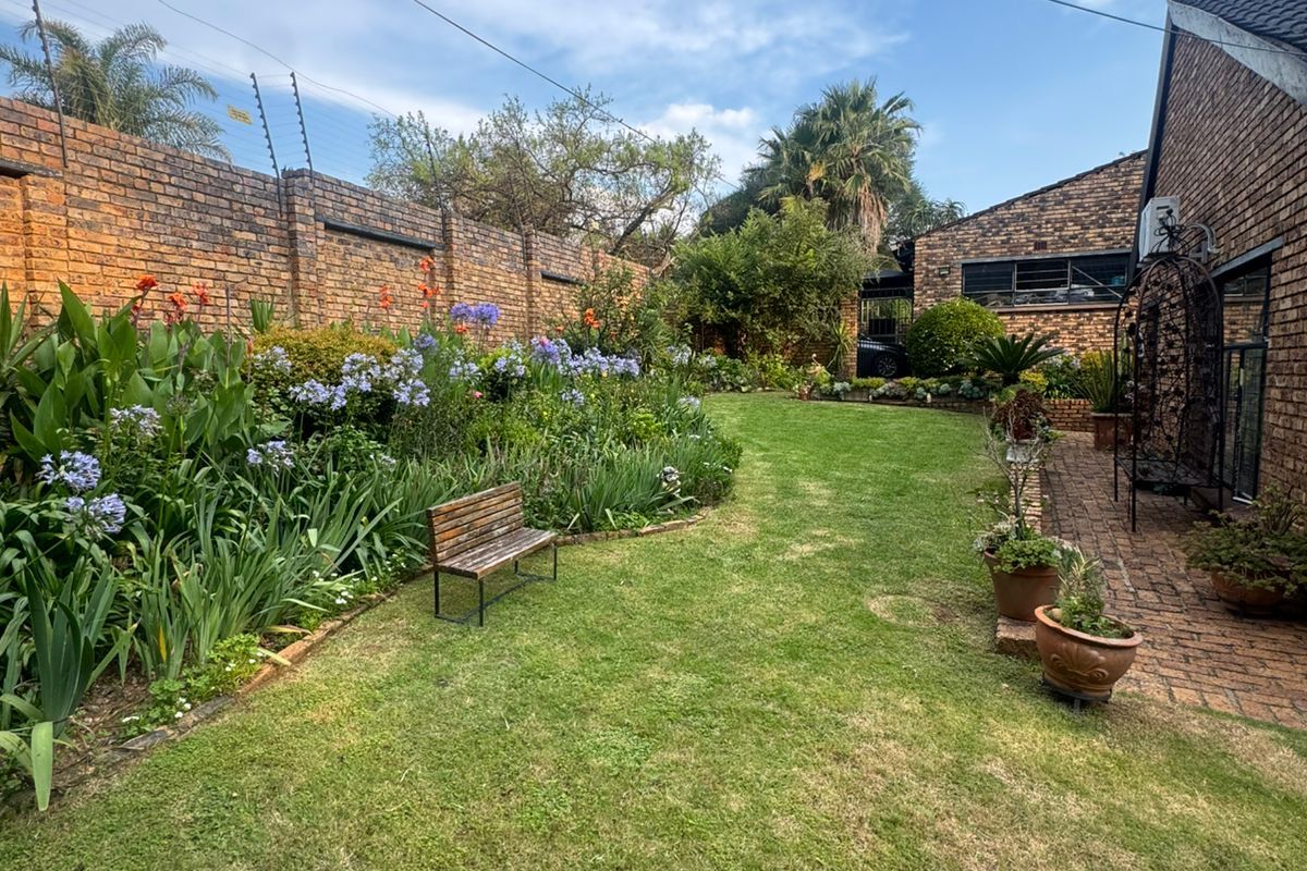 A well-maintained private garden featuring a lush lawn, established flower beds, brick boundary walls with electric fencing, and a paved side walkway.