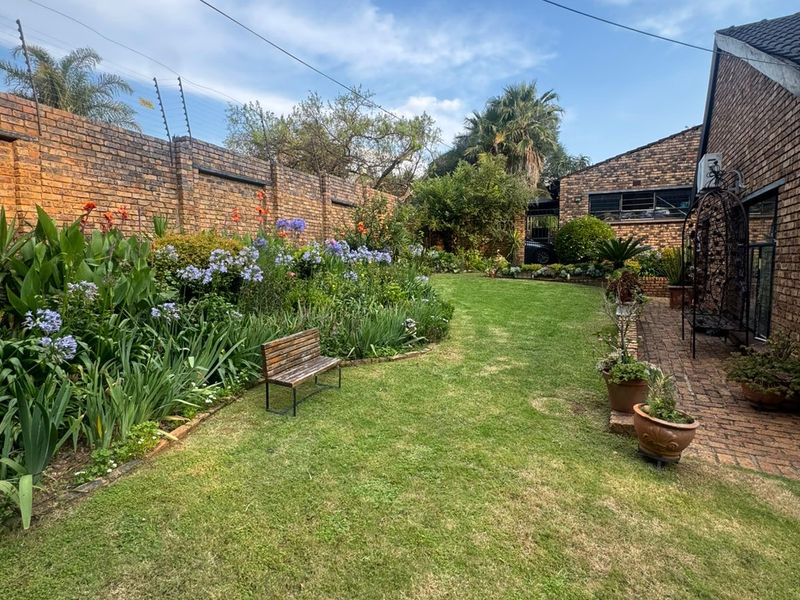 A well-maintained private garden featuring a lush lawn, established flower beds, brick boundary walls with electric fencing, and a paved side walkway.