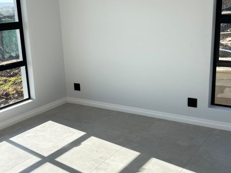 A bright, empty bedroom featuring modern grey floor tiles, crisp white walls, and black-framed windows providing natural light.
