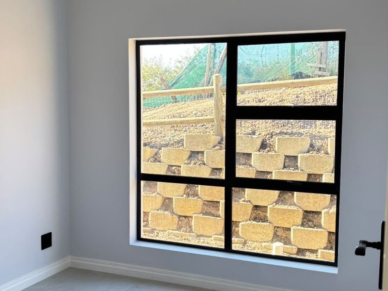 A bright, empty bedroom featuring modern grey floor tiles, recessed downlights, and a large window with a black aluminium frame overlooking a retaining wall.