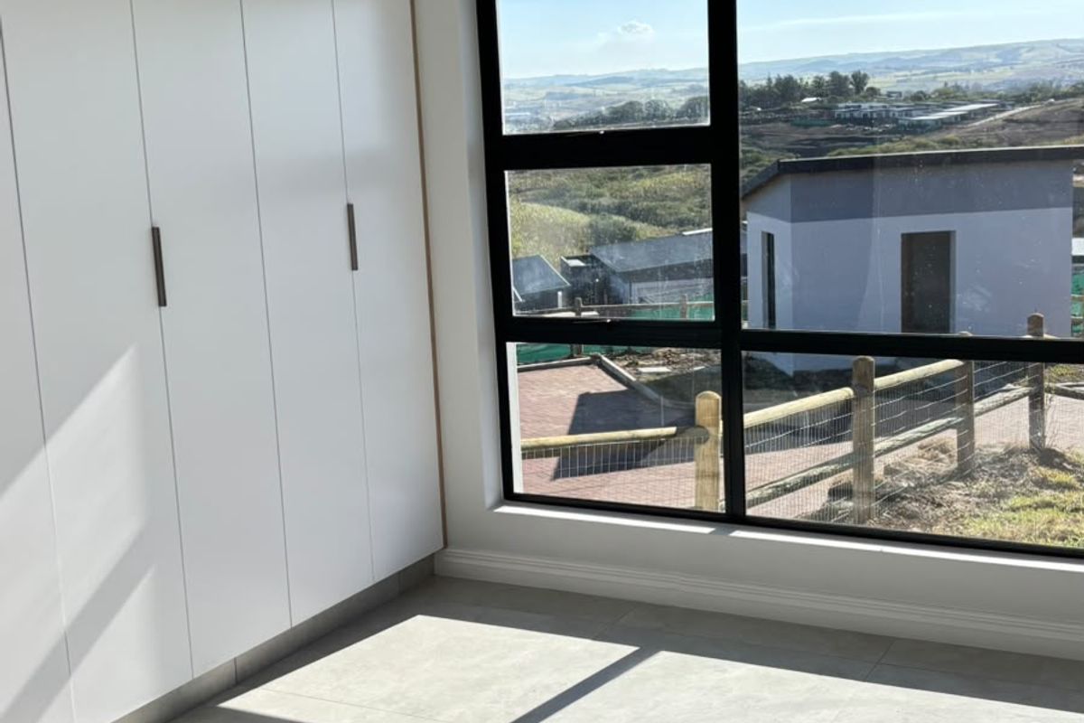 A bright, modern bedroom featuring built-in cupboards, large format floor tiles, and a large window offering scenic views.