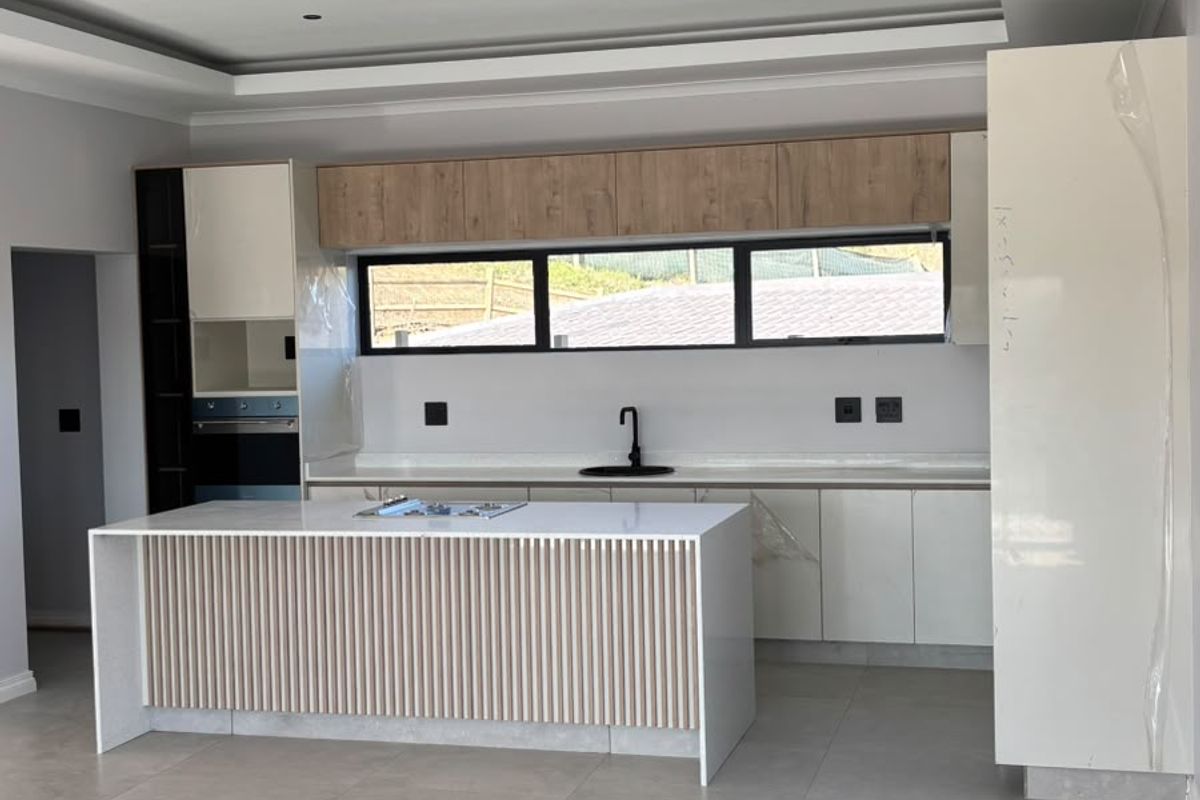 Modern, newly installed kitchen featuring a central island with slatted detailing, sleek cabinetry, built-in appliances, and neutral floor tiles in an open-plan layout.