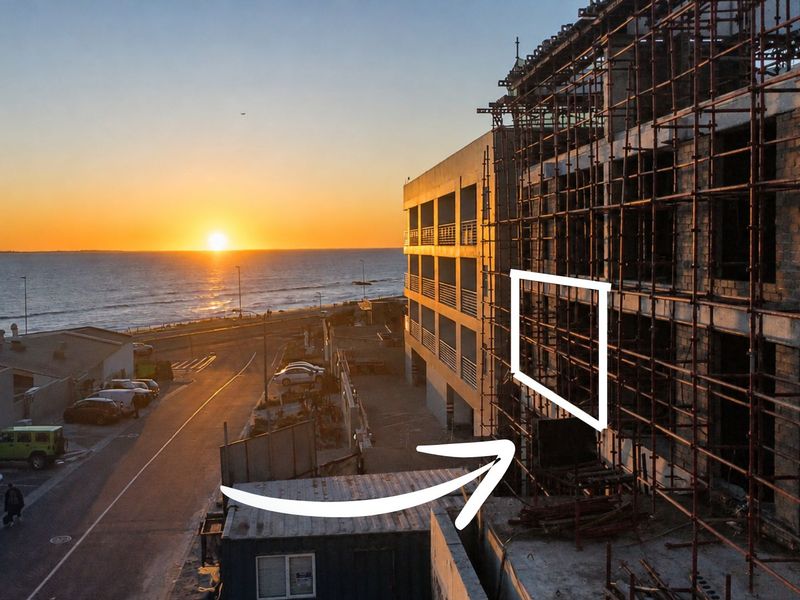 Aerial view of a multi-story apartment building under construction, featuring a concrete frame and scaffolding, situated near the coastline at sunset.