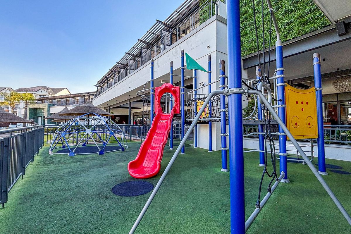 A well-maintained communal playground area featuring synthetic turf, climbing structures, and a slide, situated adjacent to a modern apartment building with covered patio space.