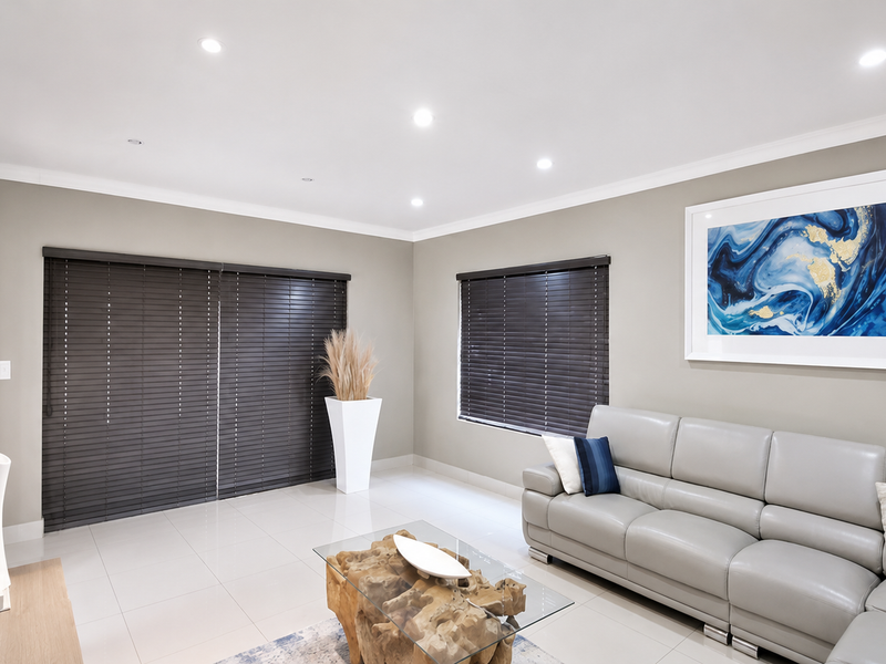 A modern, well-maintained living room featuring neutral wall tones, tiled flooring, recessed downlighting, and dark venetian blinds.