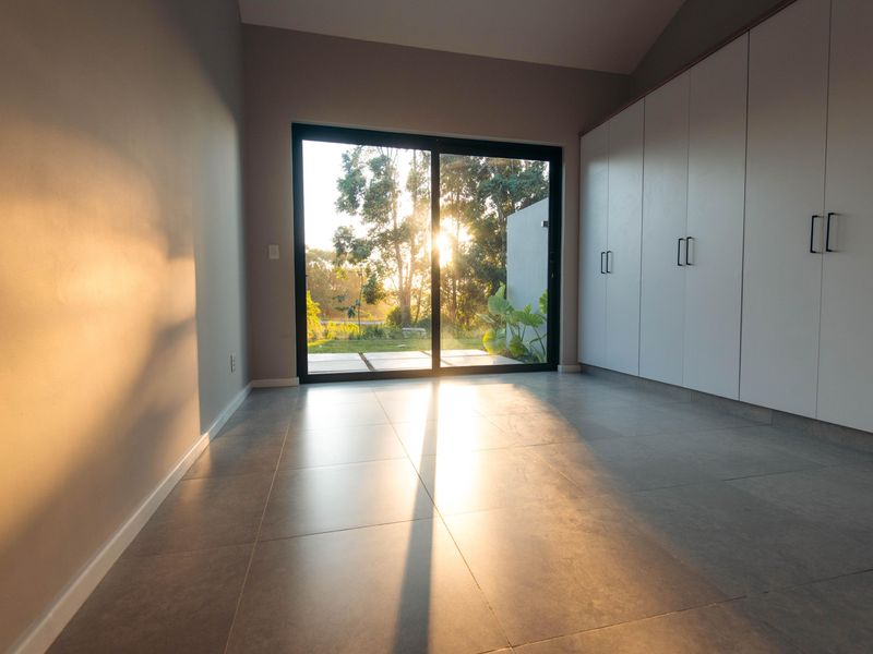 A bright, modern bedroom featuring large built-in cupboards, sleek tiled flooring, and a sliding glass door opening to a garden view.
