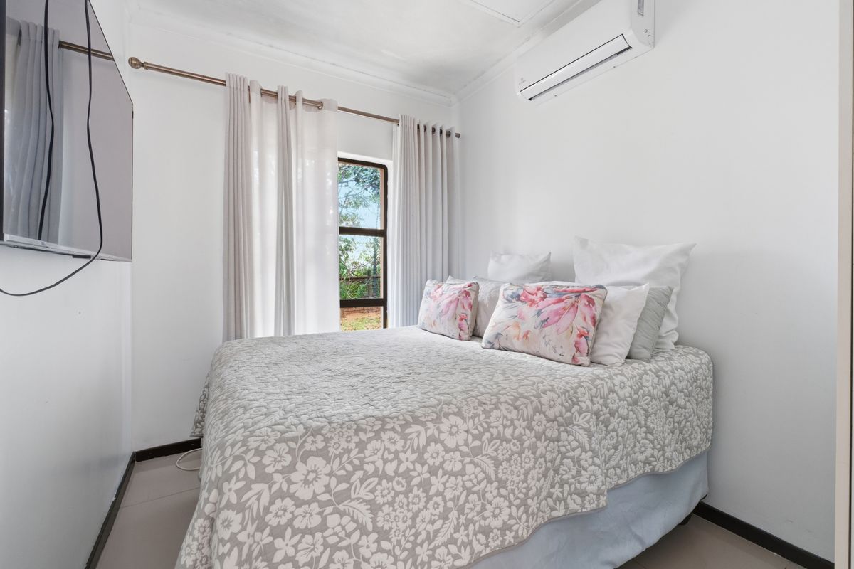 A bright and compact bedroom featuring tiled flooring, a wall-mounted air conditioning unit, and a window with curtain fixtures.