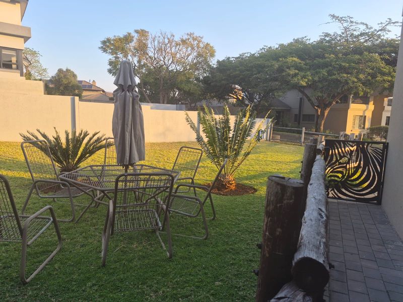 A well-maintained private garden area featuring a lawn, established cycad plants, perimeter walling, and a decorative metal gate with a paved walkway.
