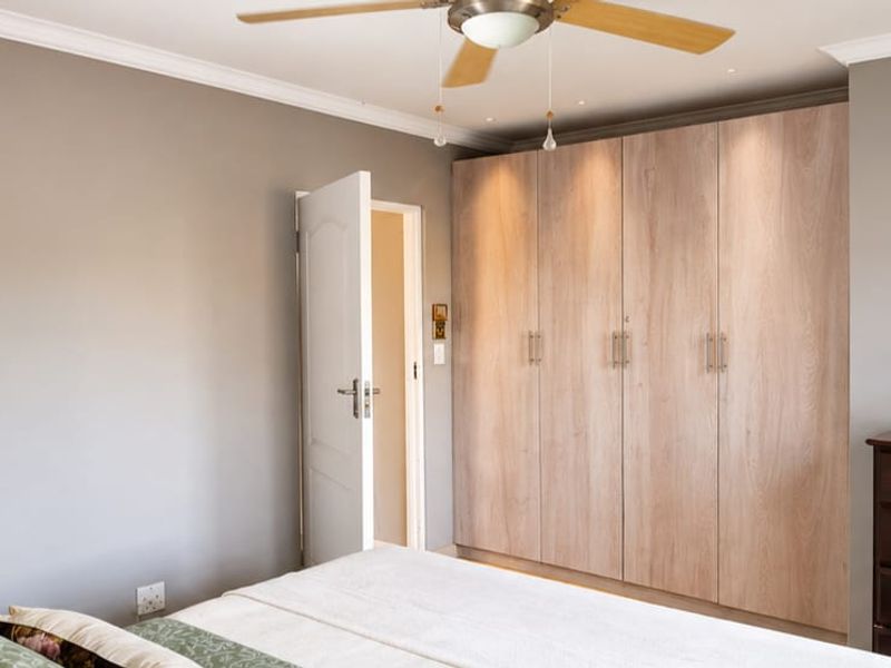 A well-maintained bedroom featuring light wood-finish built-in cupboards, a ceiling fan, and neutral grey walls with elegant coving.