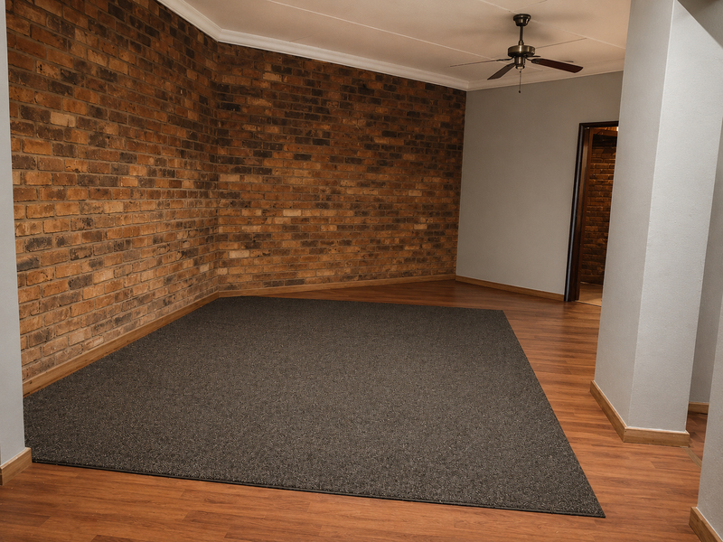 An empty living area featuring a prominent exposed brick feature wall, ceiling fan, and a combination of laminate flooring with a central carpeted section.