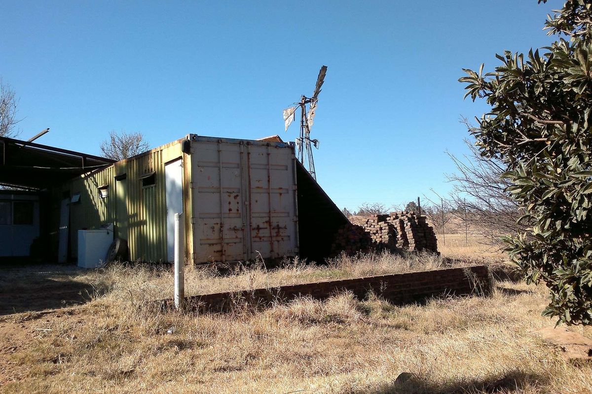 Exterior view of a farm property featuring a weathered shipping container structure, a traditional windmill, and a low brick retaining wall.