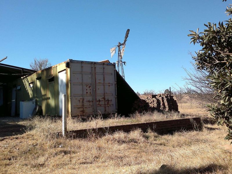 Exterior view of a farm property featuring a weathered shipping container structure, a traditional windmill, and a low brick retaining wall.