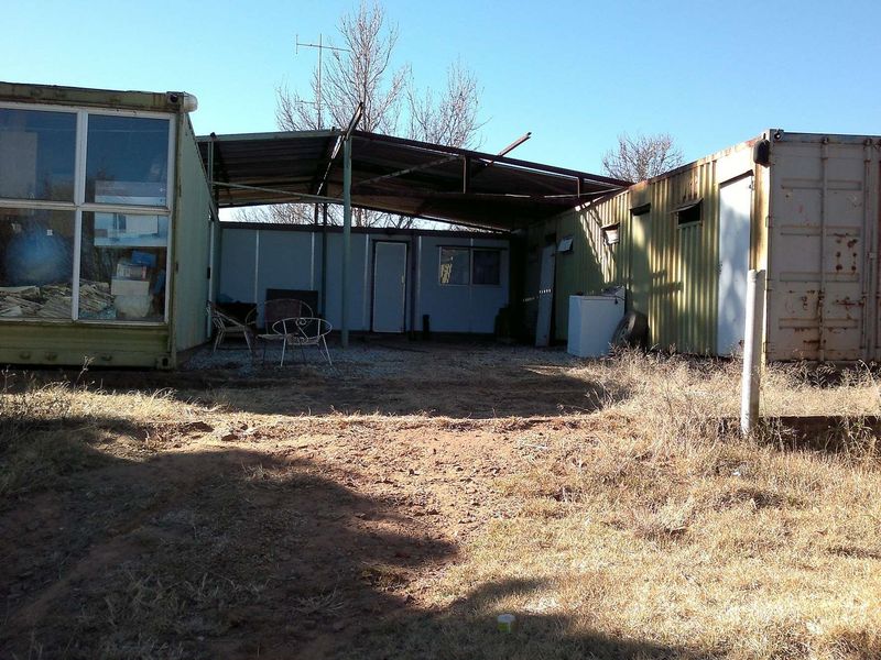 Rear exterior view of a farm property featuring shipping container structures connected by a metal roof canopy over a gravel courtyard area.