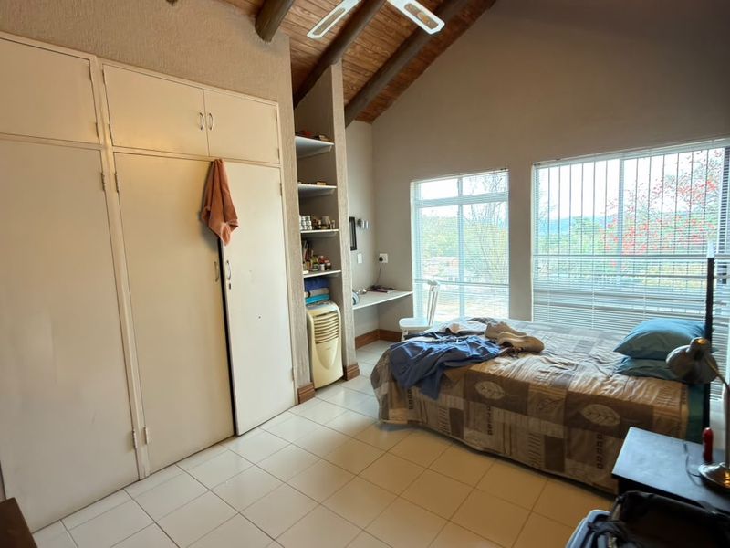 A bedroom featuring high exposed timber beam ceilings, tiled flooring, and extensive built-in cupboard storage with natural light from large windows.