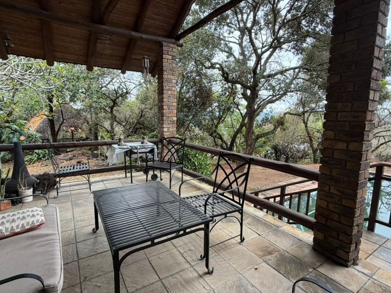 Covered outdoor patio featuring exposed timber beams, brick columns, and tiled flooring, overlooking a garden and pool area.