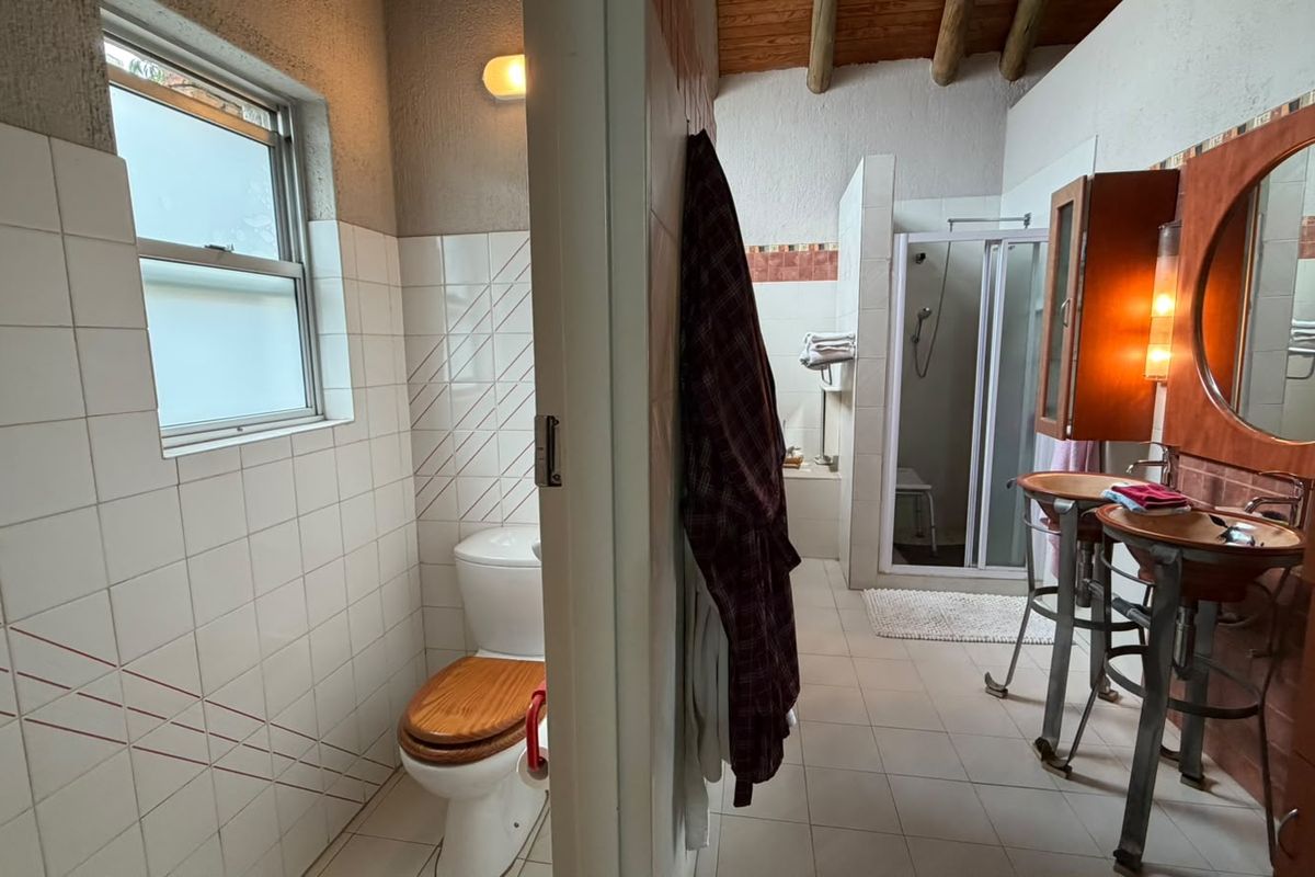 A functional bathroom featuring a separate toilet area, dual vessel sinks, a shower enclosure, and exposed timber ceiling beams with dated finishes.