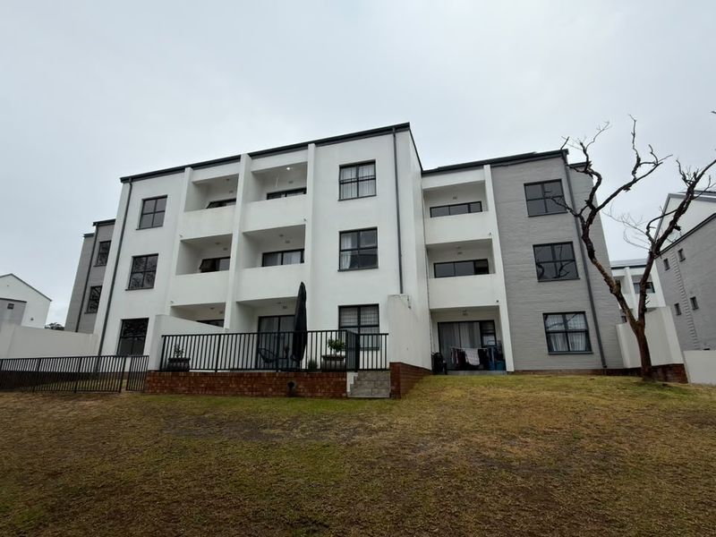 Rear view of a multi-story apartment complex featuring private balconies, ground-floor patios with metal railings, and a shared lawn area.