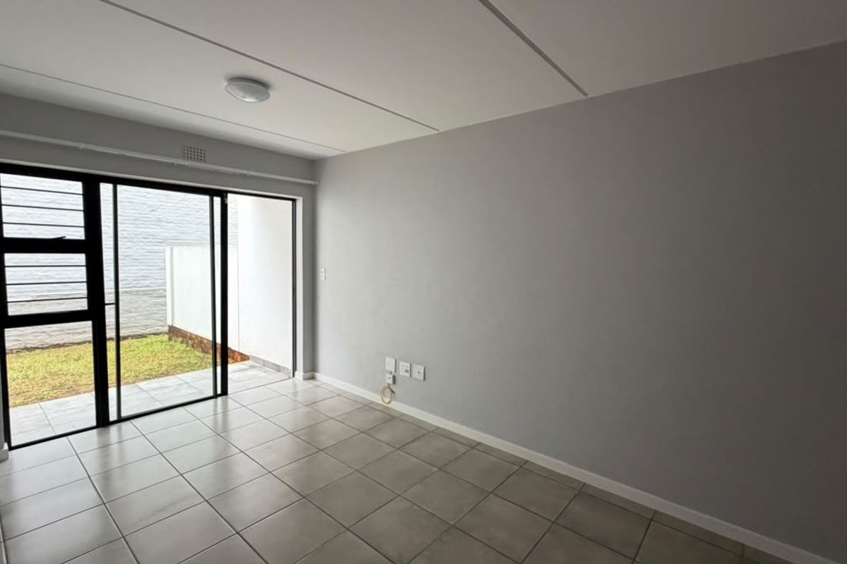 An empty living room featuring neutral tiled flooring, grey walls, and a sliding glass door providing access to a small private outdoor area.