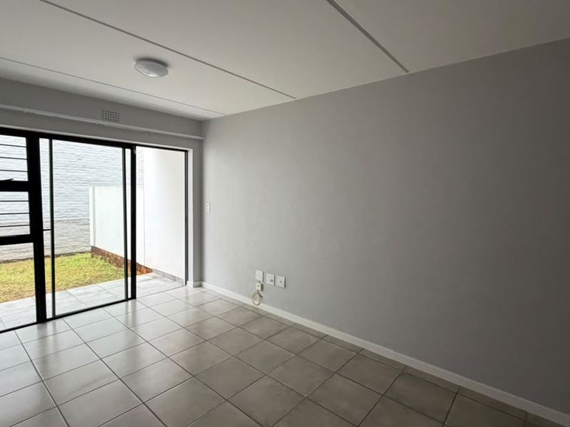An empty living room featuring neutral tiled flooring, grey walls, and a sliding glass door providing access to a small private outdoor area.