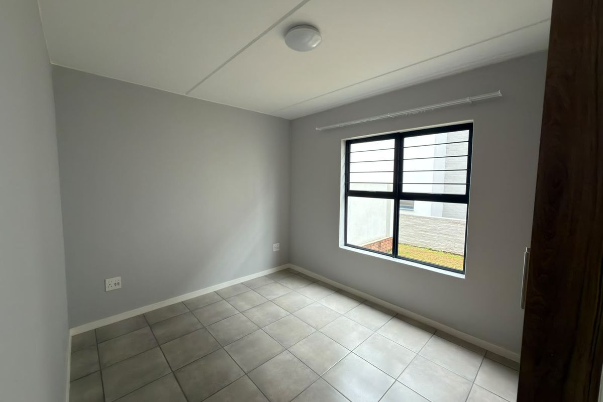A clean, empty bedroom featuring neutral grey walls, tiled flooring, and an aluminium-framed window with security bars.