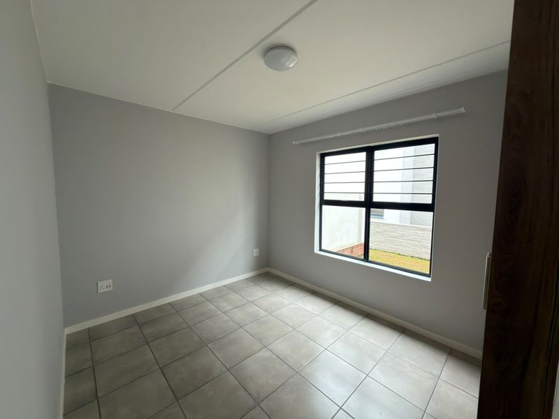 A clean, empty bedroom featuring neutral grey walls, tiled flooring, and an aluminium-framed window with security bars.