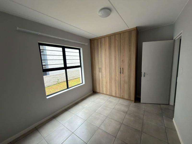 A clean, empty bedroom featuring neutral tiled flooring, a modern wood-finish built-in wardrobe, and an aluminium-framed window with security bars.