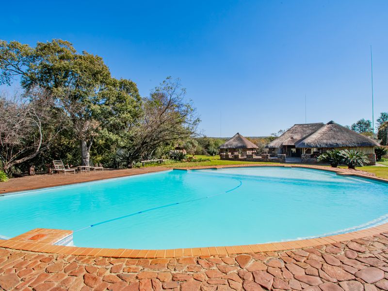 A spacious swimming pool with stone paving and wooden decking, set against a backdrop of traditional thatched-roof farm buildings and mature trees.
