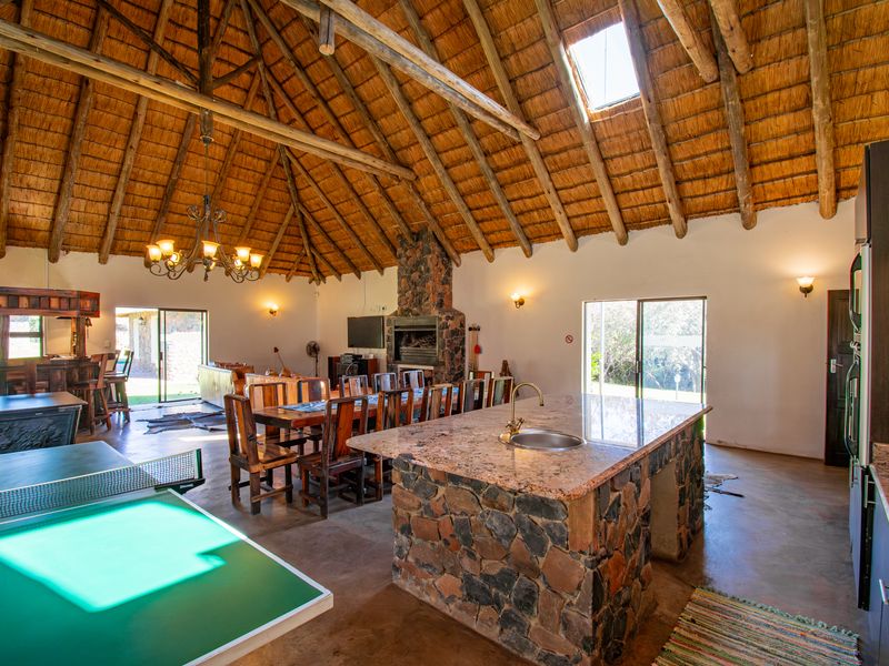 Spacious open-plan living area featuring a high thatched roof with exposed timber trusses, a stone-clad kitchen island, and a central stone fireplace.
