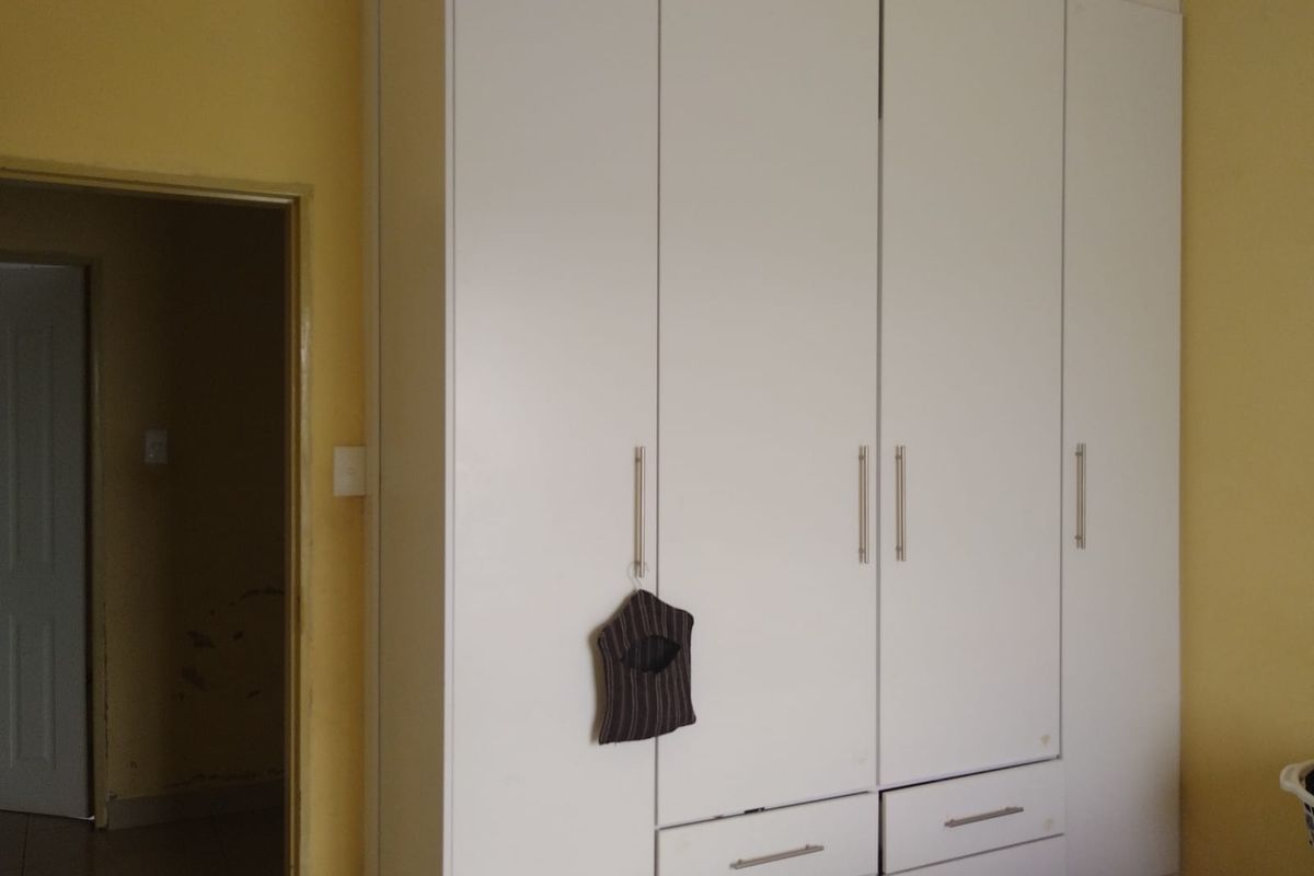 A bedroom featuring a large white built-in wardrobe with integrated drawers and metal handles, showing some visible wear on the surrounding wall surfaces.