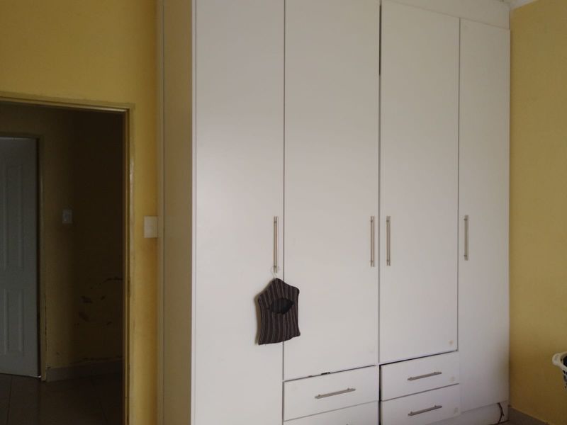 A bedroom featuring a large white built-in wardrobe with integrated drawers and metal handles, showing some visible wear on the surrounding wall surfaces.