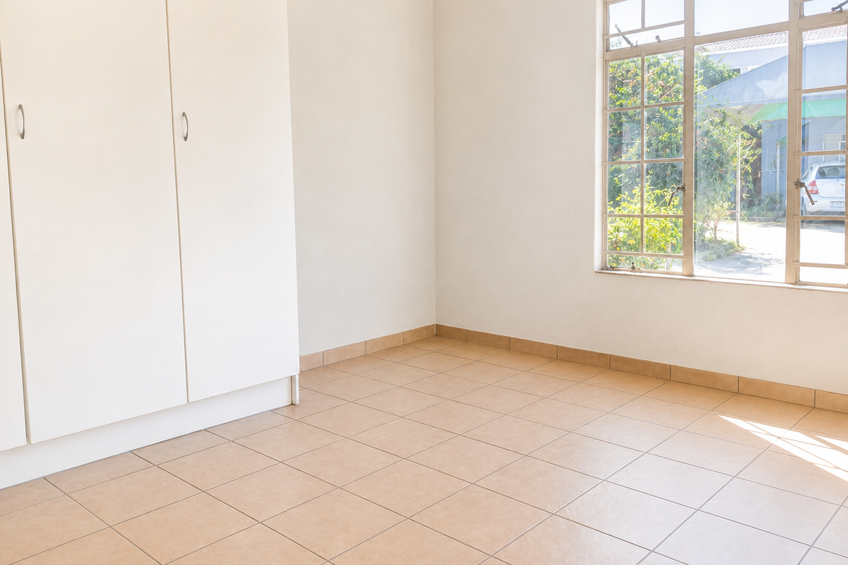 A bright, empty bedroom featuring neutral tiled flooring, built-in cupboards, and a large window providing natural light and a view of the exterior.
