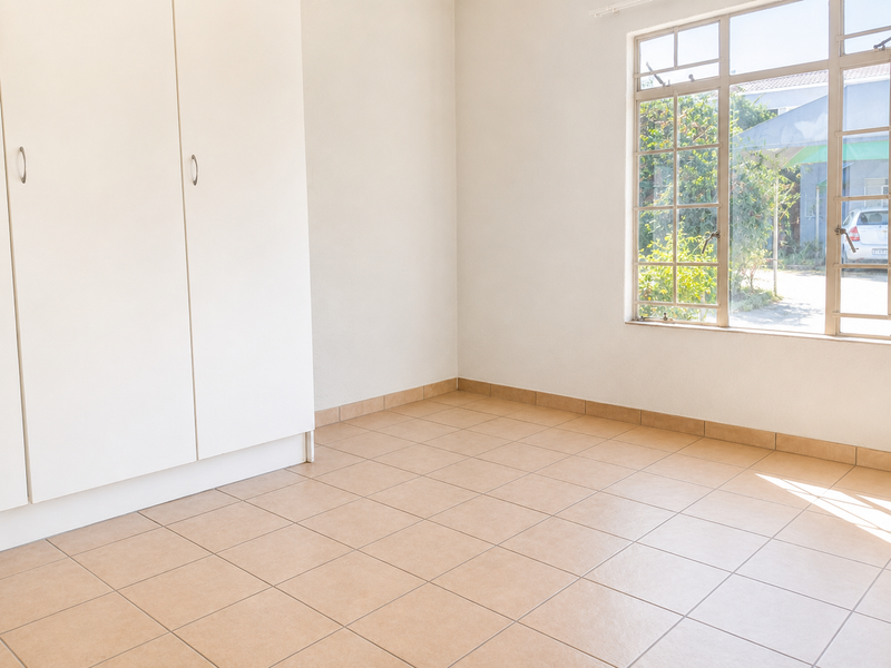 A bright, empty bedroom featuring neutral tiled flooring, built-in cupboards, and a large window providing natural light and a view of the exterior.