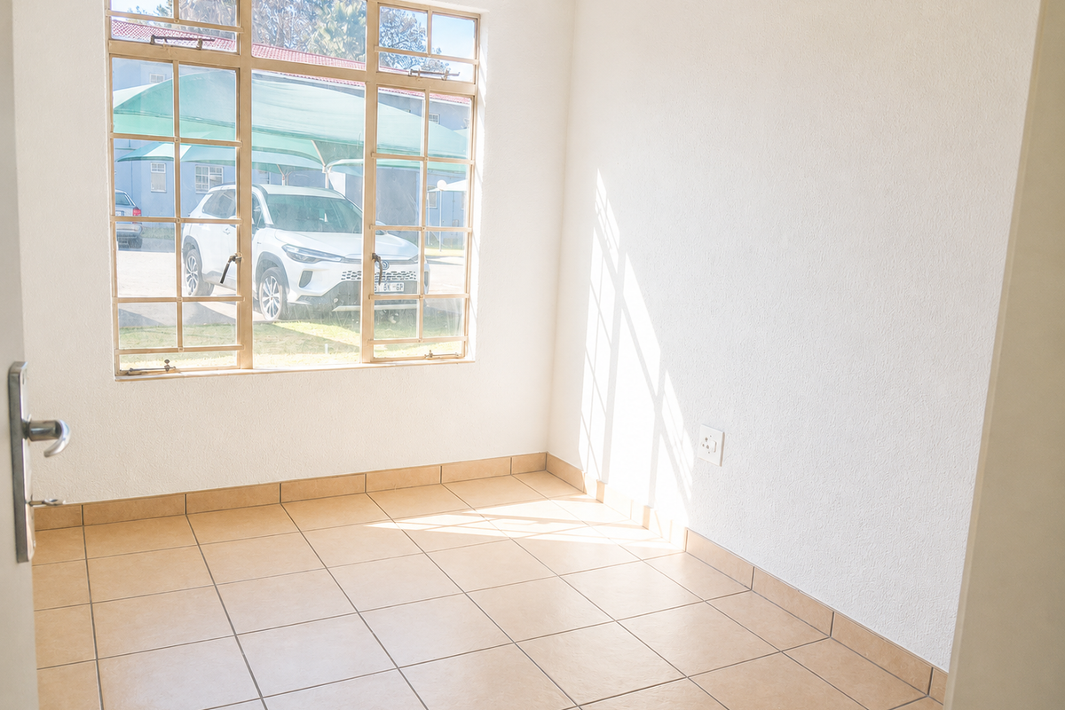 A bright, empty living area featuring neutral tiled flooring and a large window providing natural light and a view of the exterior parking area.