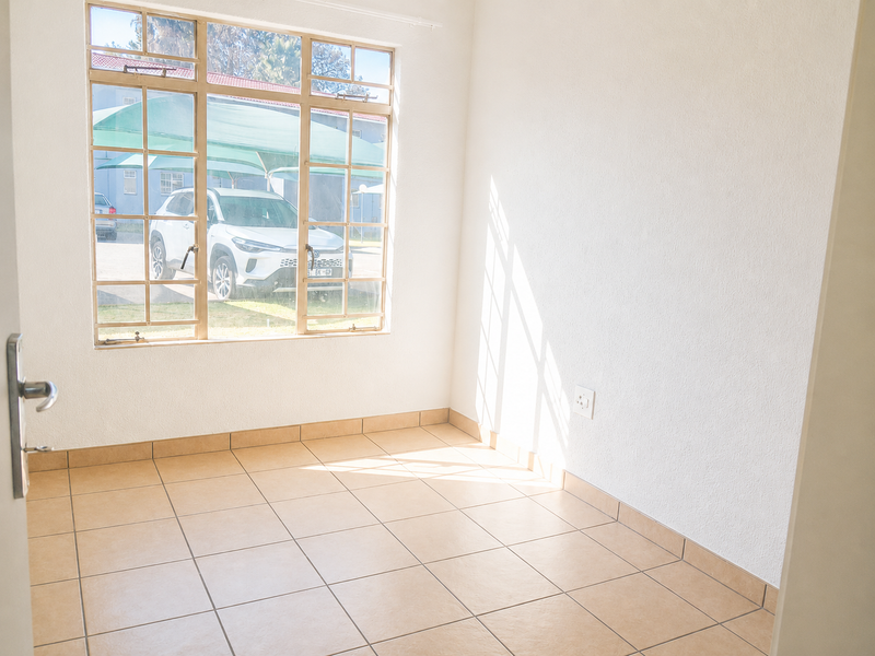 A bright, empty living area featuring neutral tiled flooring and a large window providing natural light and a view of the exterior parking area.