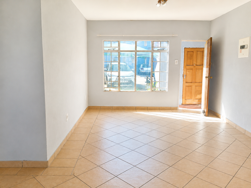 A bright, empty living area featuring neutral tiled flooring, a large multi-pane window, and a traditional timber entrance door.