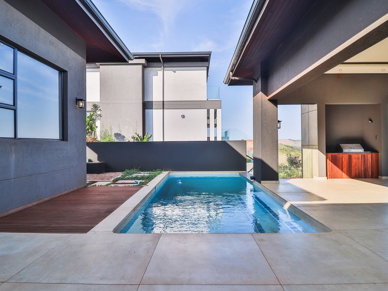 Modern outdoor entertainment area featuring a swimming pool, covered patio with built-in braai, and timber decking, set against a contemporary architectural backdrop.