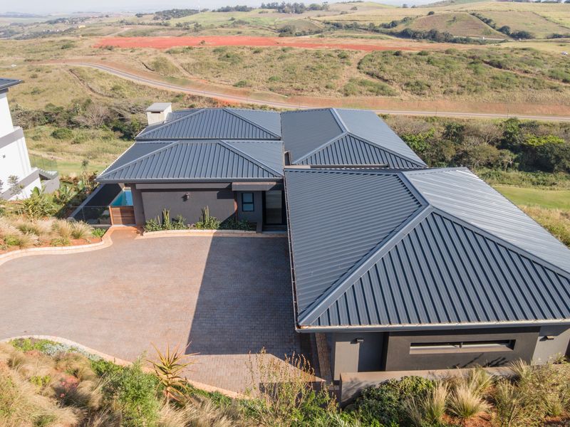 Aerial view of a modern house featuring a dark metal roof, paved driveway, and a swimming pool, set against a scenic landscape.
