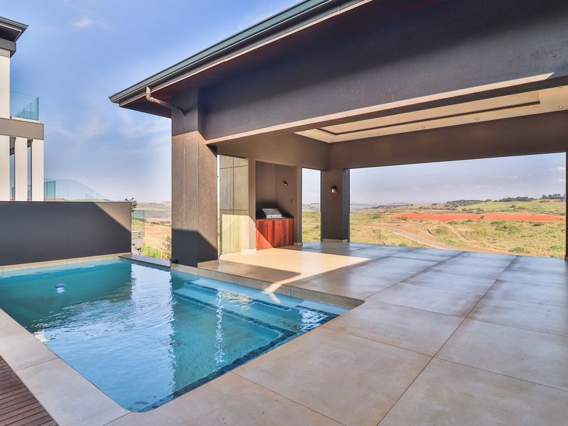 A modern covered patio area featuring a built-in braai and a sparkling swimming pool, overlooking expansive scenic landscape views.