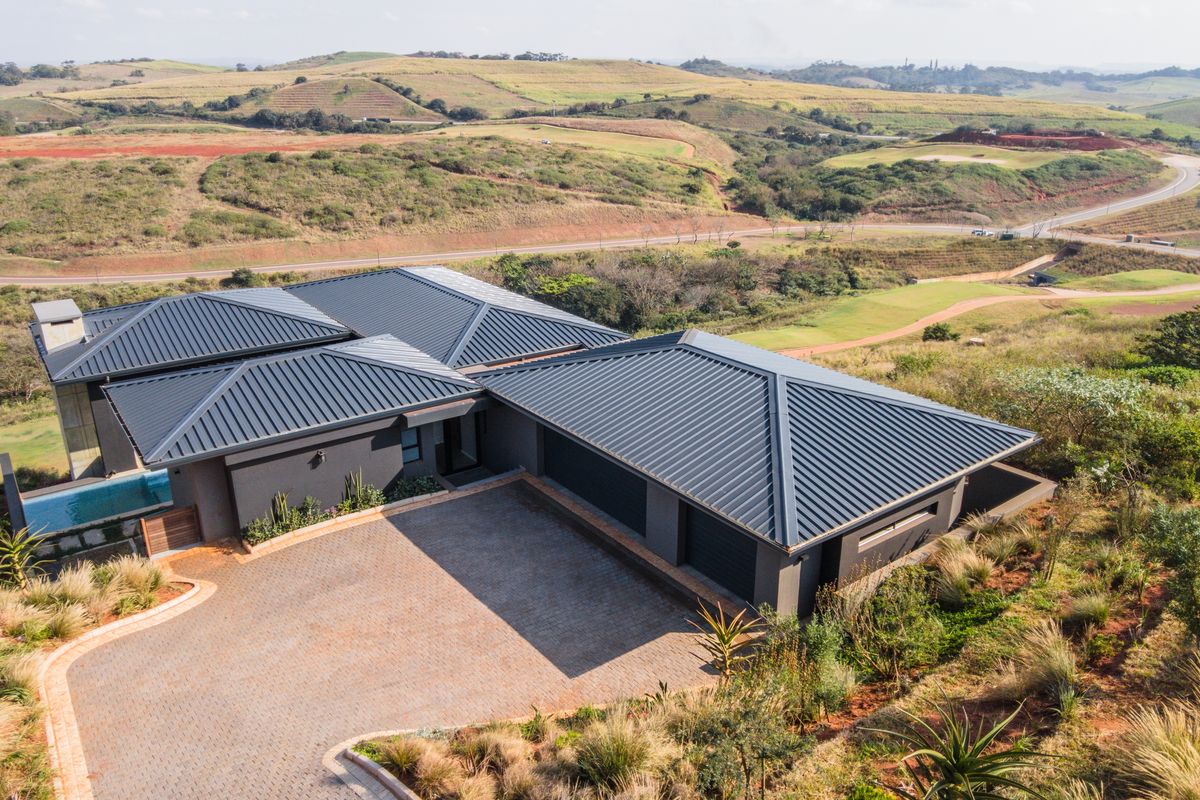Aerial view of a modern house featuring a dark metal roof, paved driveway, double garage, and a swimming pool set in a scenic landscape.