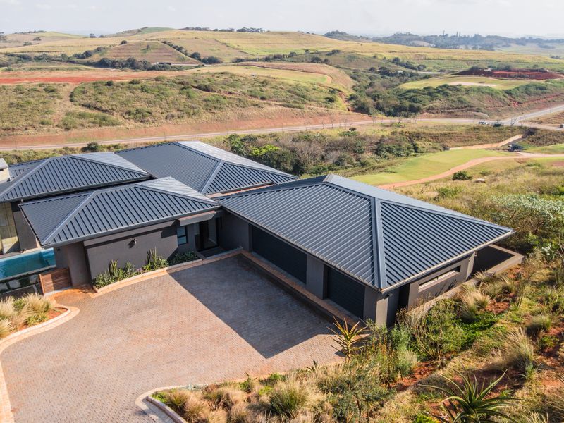 Aerial view of a modern house featuring a dark metal roof, paved driveway, double garage, and a swimming pool set in a scenic landscape.