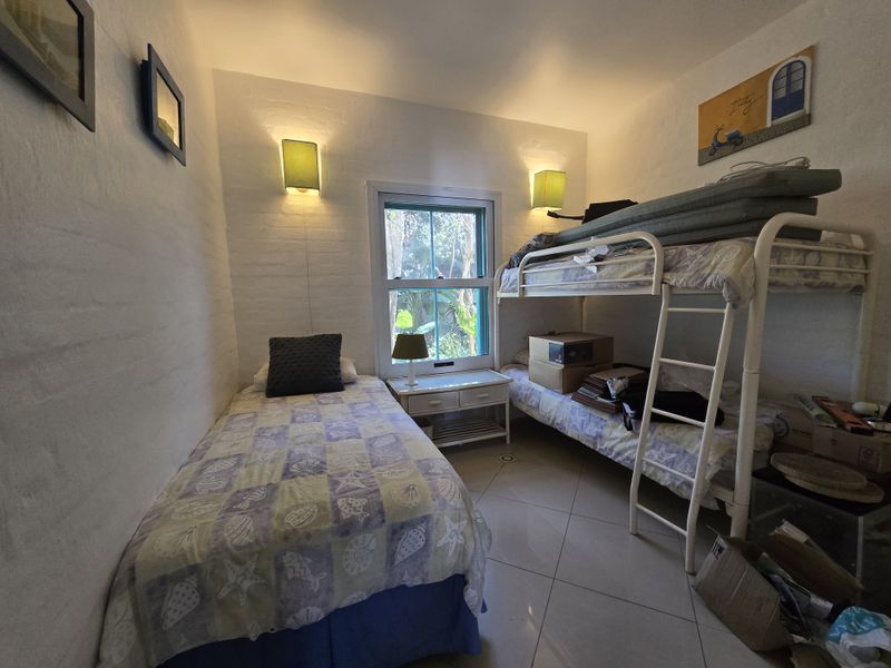 A compact bedroom featuring tiled flooring, wall-mounted lighting, and a sash window overlooking greenery, currently configured with multiple sleeping arrangements.