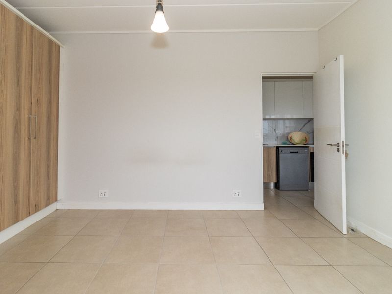 An empty living area featuring neutral tiled flooring and built-in wooden cupboards, with a doorway leading to a kitchen space.