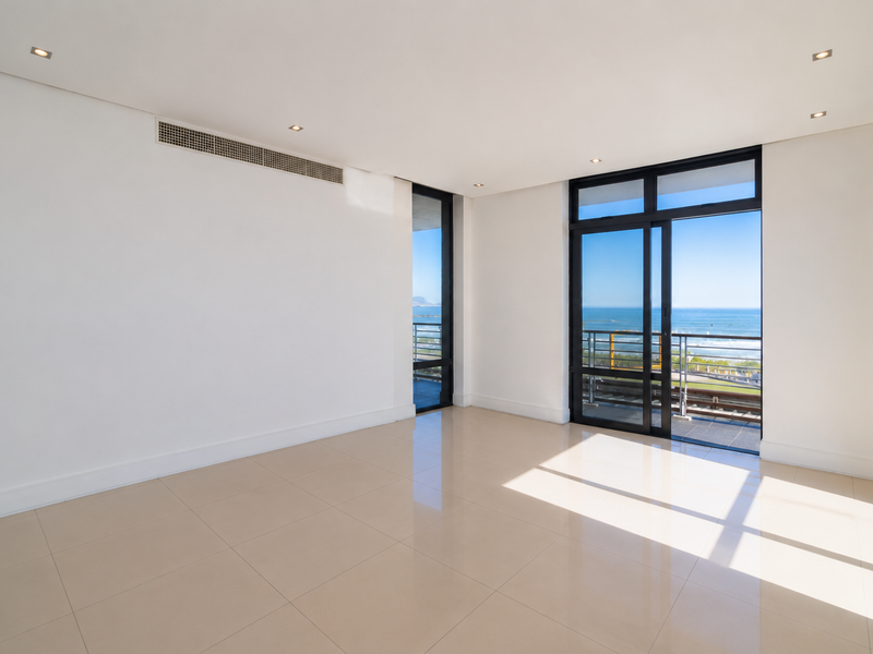 A bright, spacious, and empty living area featuring polished tile floors, recessed lighting, and large sliding glass doors opening to a scenic ocean view.